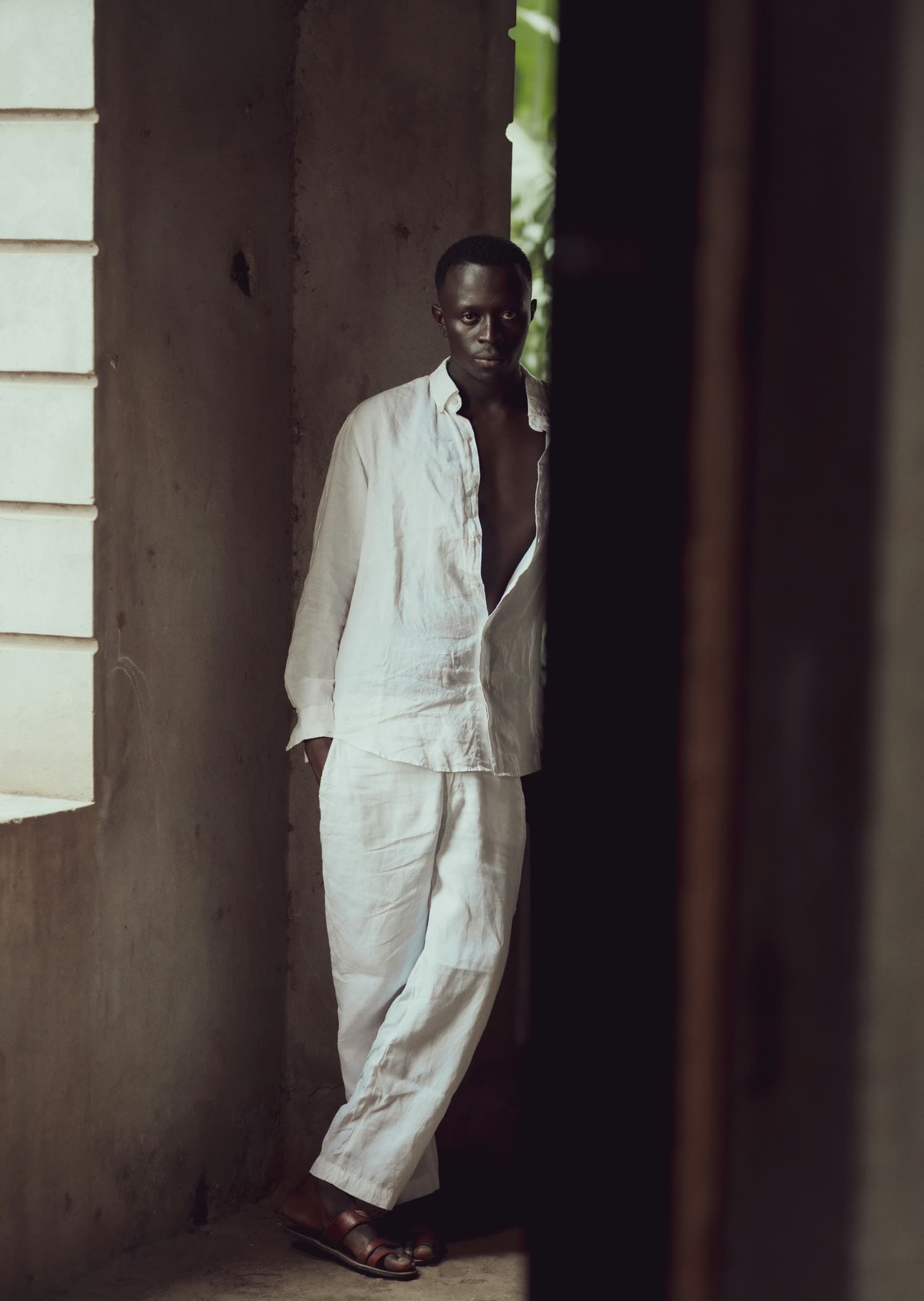 The undyed linen shirt worn open with matching trousers, leaning in a doorway against bare concrete